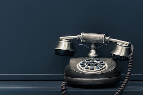 An old fashioned blacked corded telephone against a dark blue wall