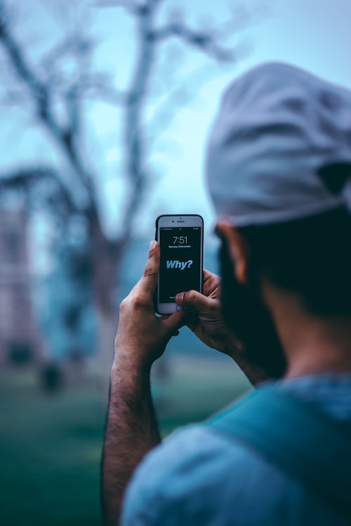 A man holding his iPhone out to take a picture with the word why displayed on the screen